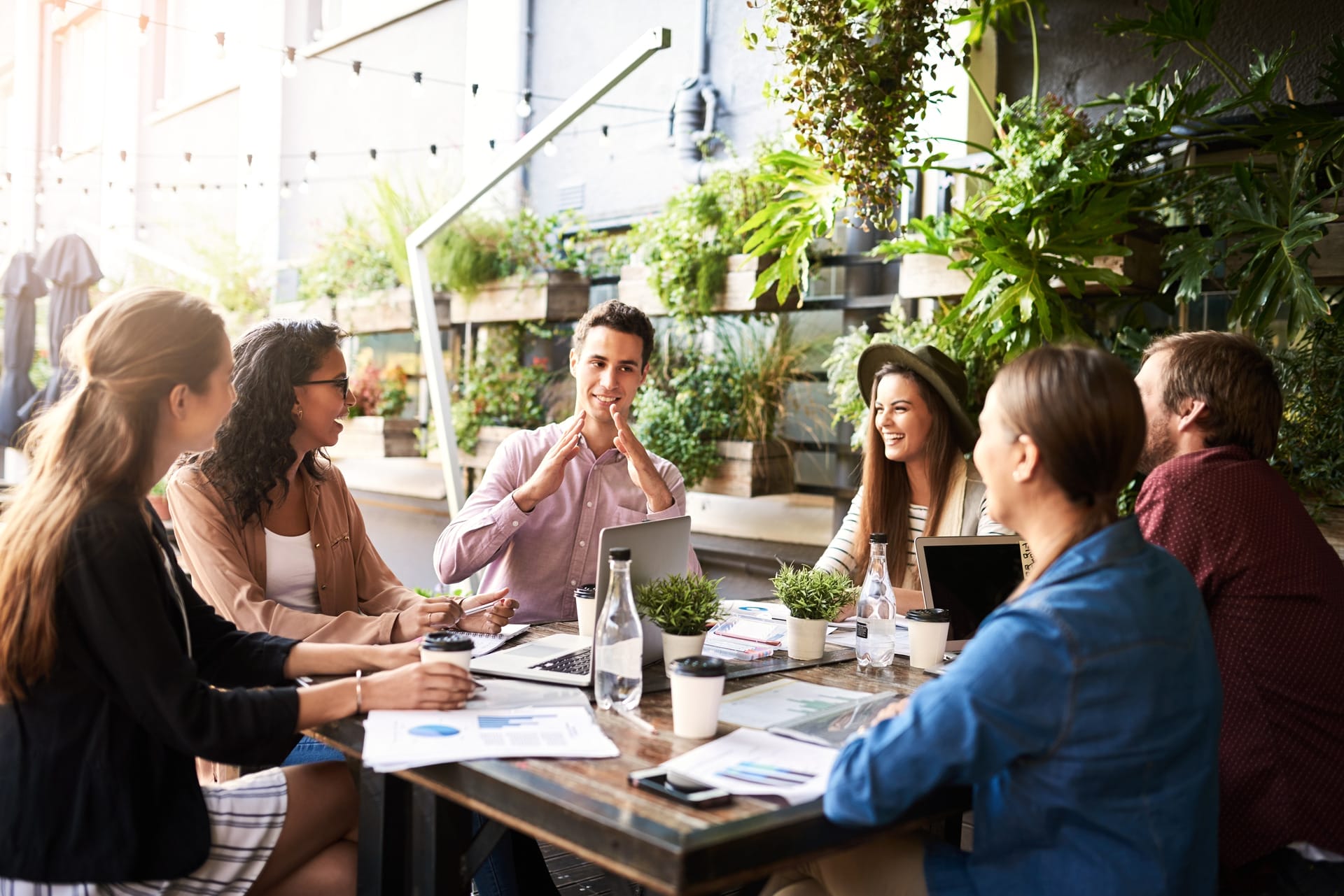 Groupe de six personnes assises autour d’une table, avec ordinateurs portables, documents et tasses de café, échangeant dans un espace lumineux décoré de plantes.